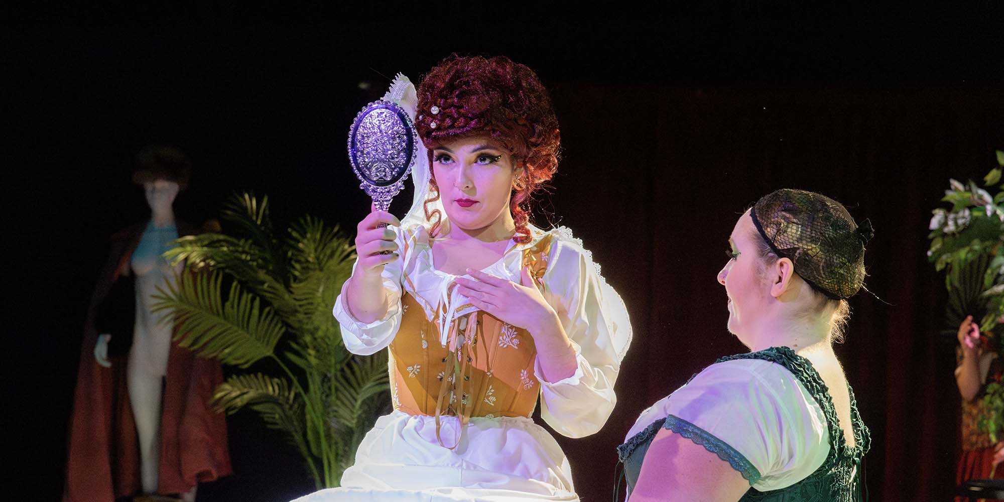 An actor wearing a bright red wig with pearls, large Elizabethan ruff, gold corset, and farthingale admires herself in a hand mirror while an actor in a green corset kneels at their feet.