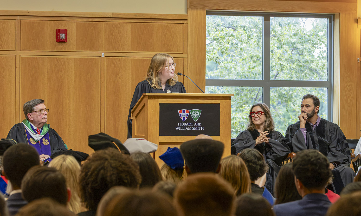 HWS Student Government President Ela Gardiner '27 speaks during Convocation, while President Mark D. Gearan, former trustee Jane Erickson '07 and Trustee Paul Wasmund '07 look on.   