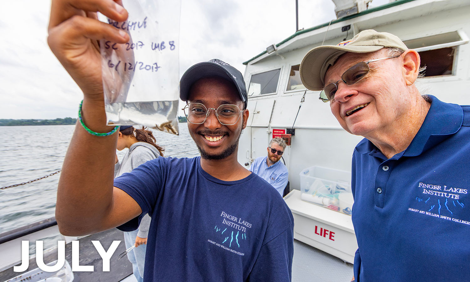 July means summer research aboard the William Scandling. Here, Finger Lakes Institute research student Daudi Aden ’26 shows Daniel R. Baker ’72 organisms collected from sediment in Seneca Lake.  
