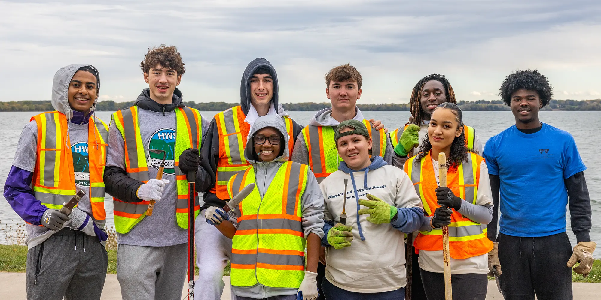 Students in high-visibility safety vests holding rakes and shovels on the shore of Seneca Lake during the HWS Day of Service