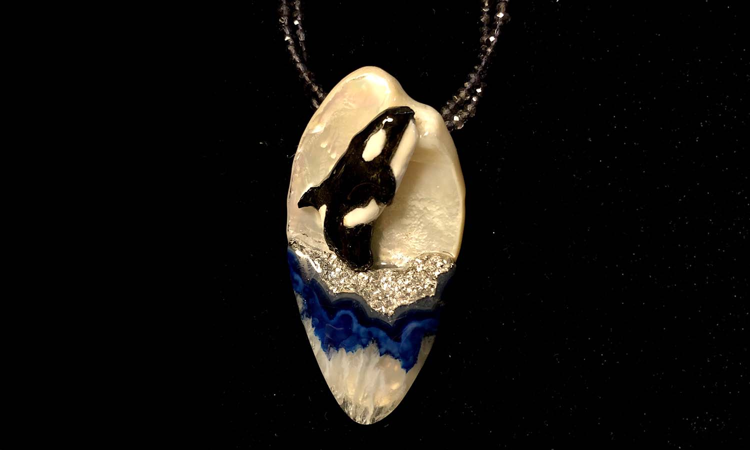 "Breaching Orca" is carved in jet with warthog white accents, mounted on a golden pearl shell, above a sea of platinum Oco agate.